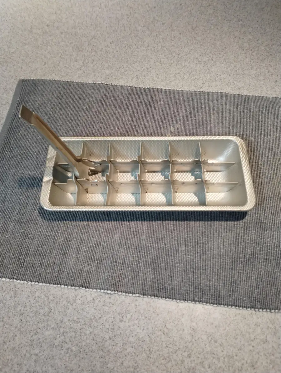 A vintage metal ice cube tray with a lever sits on a gray textured mat on a countertop.