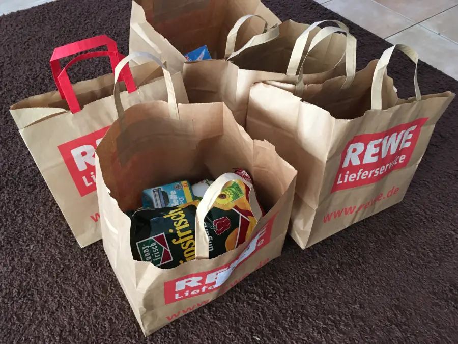 Four brown paper shopping bags with “REWE Lieferservice” branding sit on a brown carpet, filled with various groceries, including chips and packaged food items.