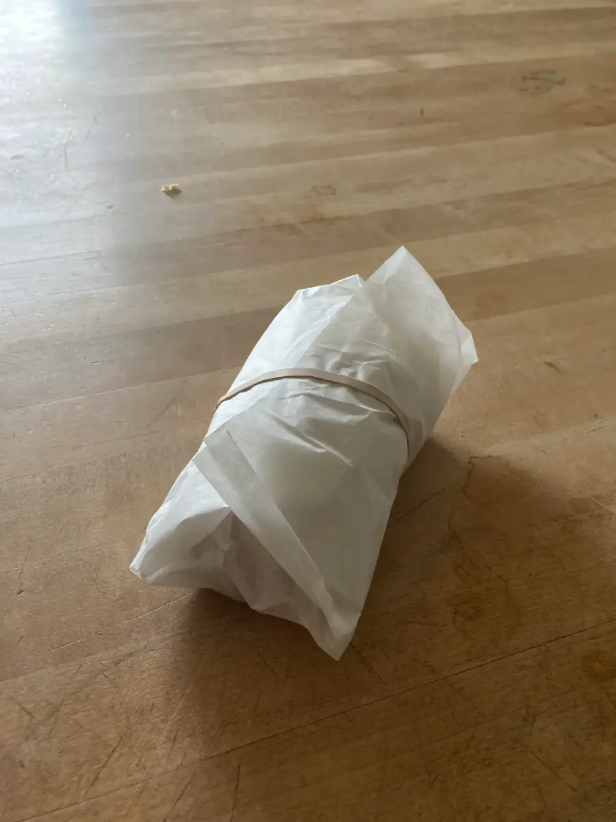 A small object wrapped in white parchment paper and secured with a rubber band sits on a wooden surface.