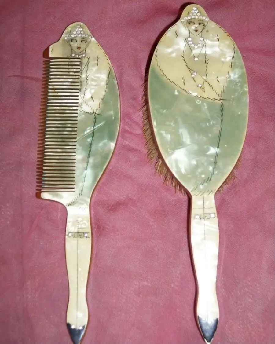 A vintage comb and brush set with handles decorated to resemble women in elegant dresses, set on a pink fabric background.