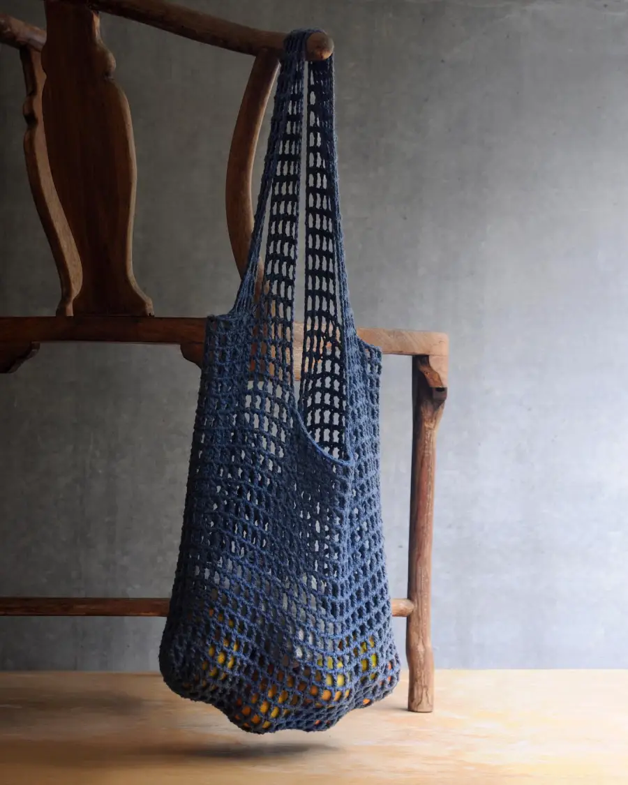 A blue crocheted mesh bag hangs from a wooden chair with a grey wall in the background. There are some indistinct items inside the bag, possibly fruits or vegetables.