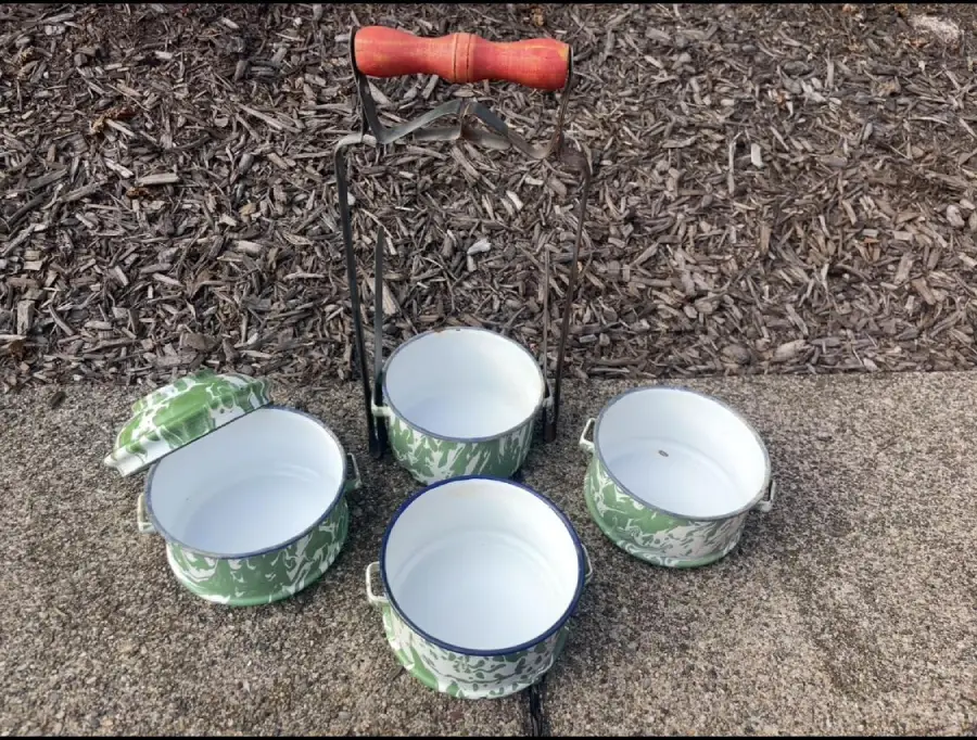 Three green-and-white enamel pots with handles and lids, arranged on the ground with a metal carrying rack featuring a red handle, placed on a concrete and mulch surface.