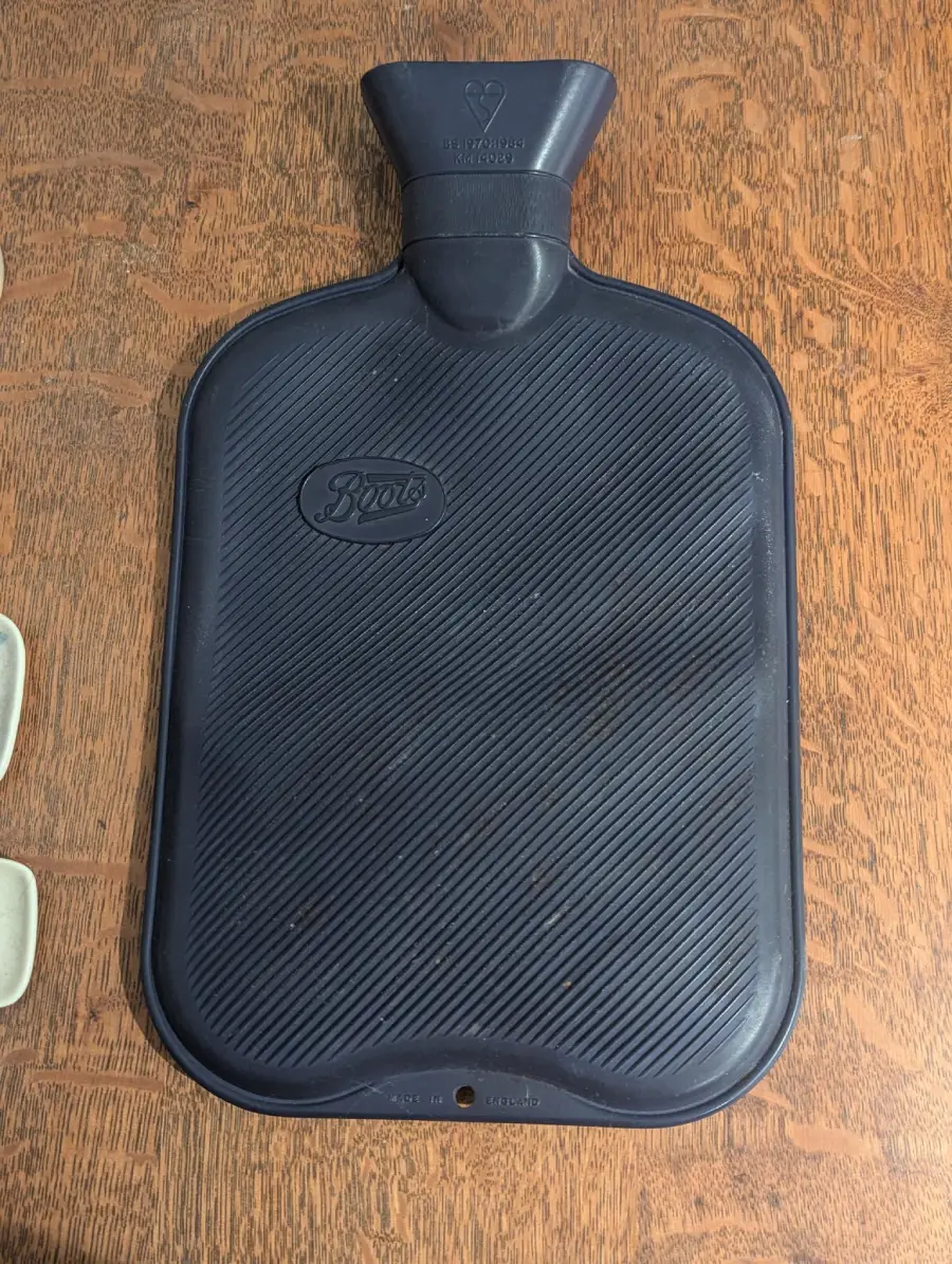 A black rubber hot water bottle with a textured surface and a capped opening at the top, placed on a wooden table. The word “Boots” is embossed in the center of the bottle.