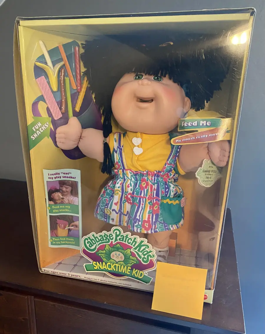 Cabbage Patch Kids Snacktime Kid doll in original box, wearing colorful overalls and holding plastic snacks. Packaging features bright colors and text promoting its interactive feeding feature.