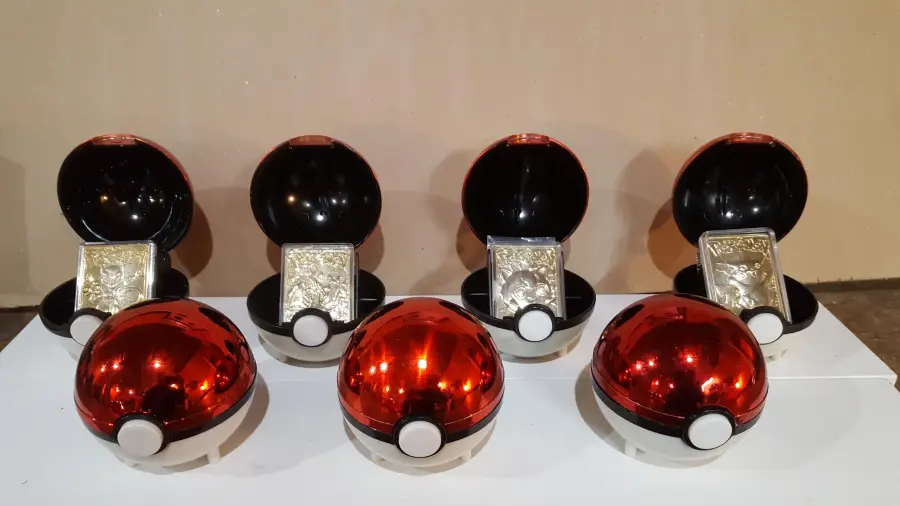 Five shiny red Poké Balls are open, each displaying a metallic Pokémon card inside. Three unopened Poké Balls are placed in front of them on a white surface, against a plain beige background.