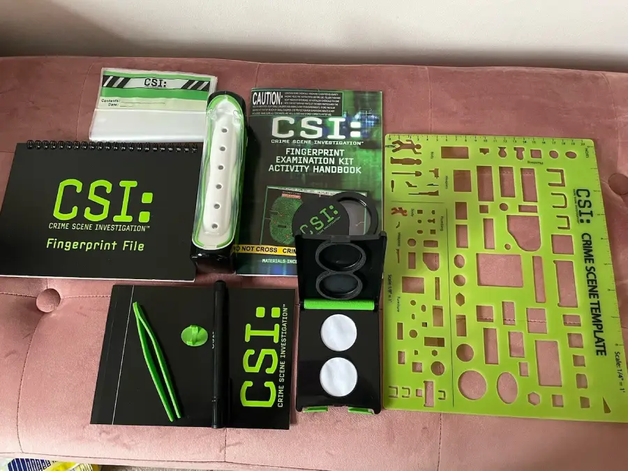CSI kit on a pink surface, including a fingerprint file, templates, brushes, powders, evidence cards, green tools, and an activity handbook for crime scene investigation activities.