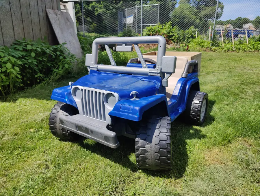 A blue toy jeep with large tires sits on green grass in a backyard, with a wooden fence, plants, and a chain-link fence in the background under a sunny sky.