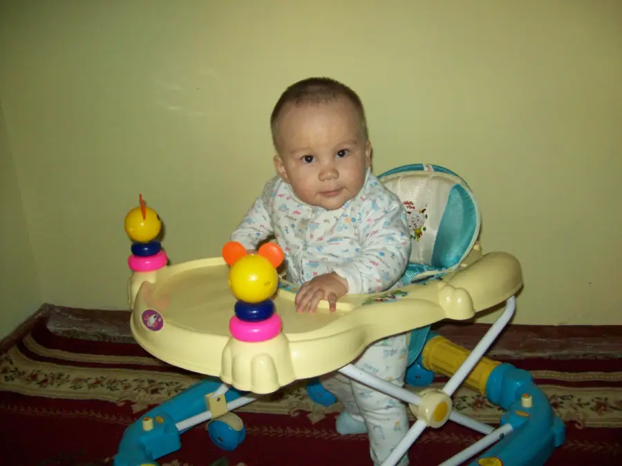 A baby in a light-patterned onesie sits in a yellow and blue baby walker, looking at the camera. The room has pale yellow walls and a red-patterned carpet.