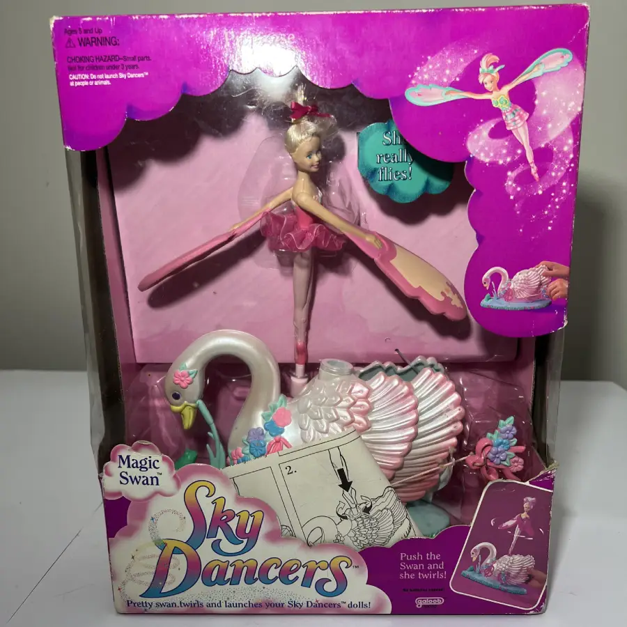 Boxed Sky Dancers Magic Swan toy with a winged fairy doll and a decorative swan launcher. Packaging is pink with illustrations and instructions, and the text “Sky Dancers Magic Swan” is visible on the box front.