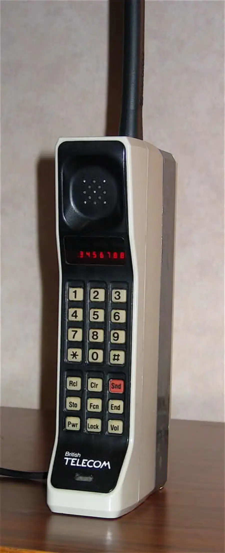 A vintage British Telecom mobile phone with a large antenna, numeric keypad, and a small digital display, sitting on a wooden surface.