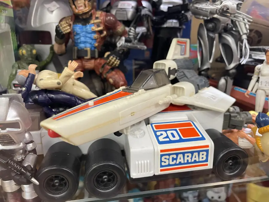 A white toy race car labeled “20 SCARAB” sits on a shelf surrounded by various action figures and robot toys, creating a colorful and nostalgic display.