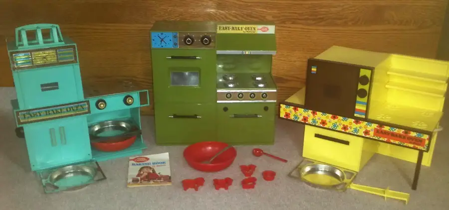 Three vintage Easy-Bake Ovens in teal, green, and yellow, surrounded by red mixing bowls, utensils, baking trays, and a recipe booklet, all set against a wooden background.