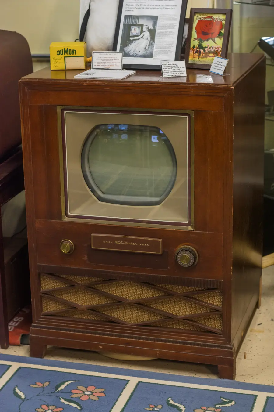 An old wooden television set with a rounded screen, speaker grill, and two knobs. The TV sits on the floor, surrounded by informational cards and vintage memorabilia.