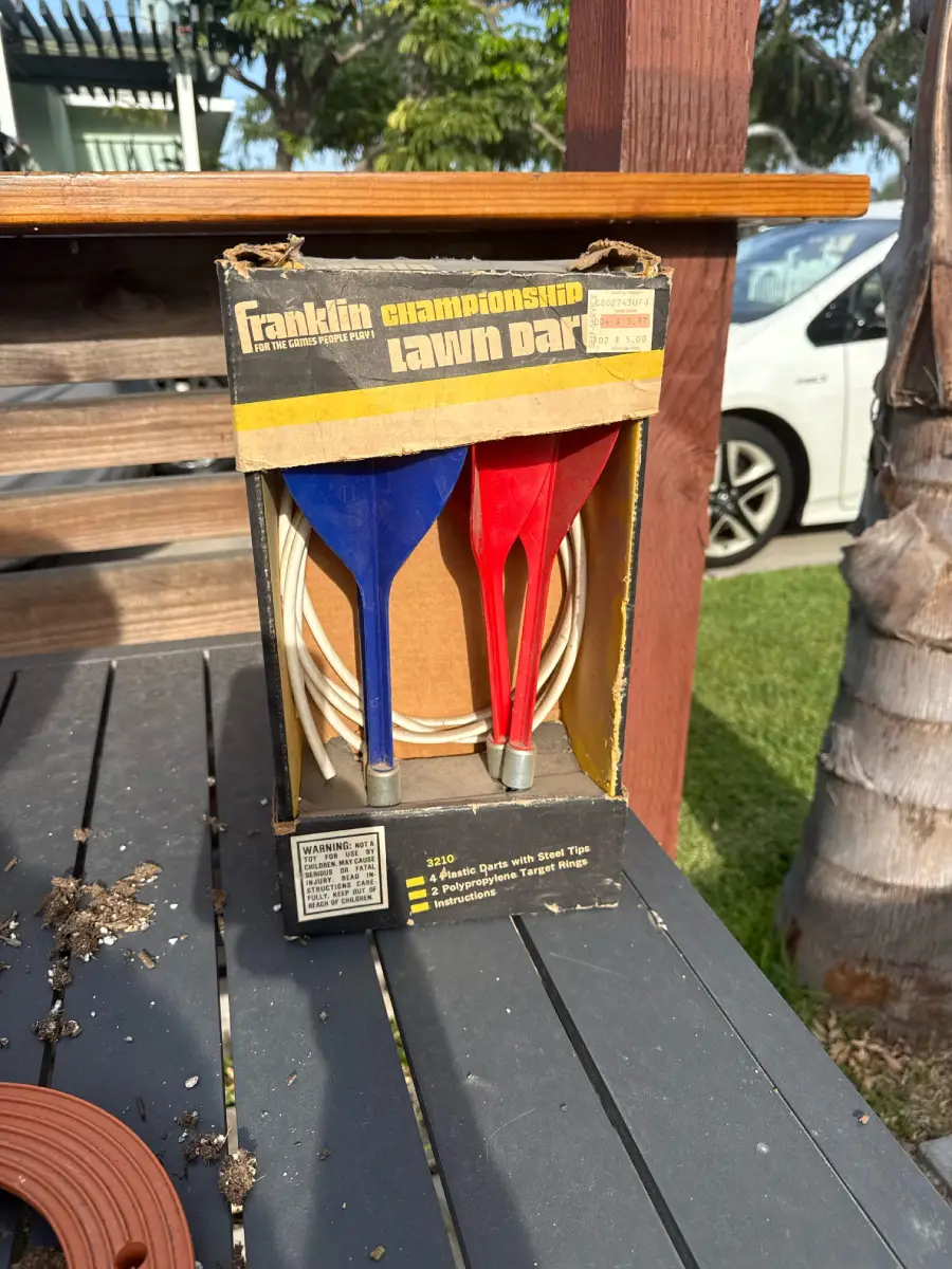 A box of Franklin championship lawn darts sits on a patio table, containing two blue and two red darts with white rings. A white car and trees are visible in the background.