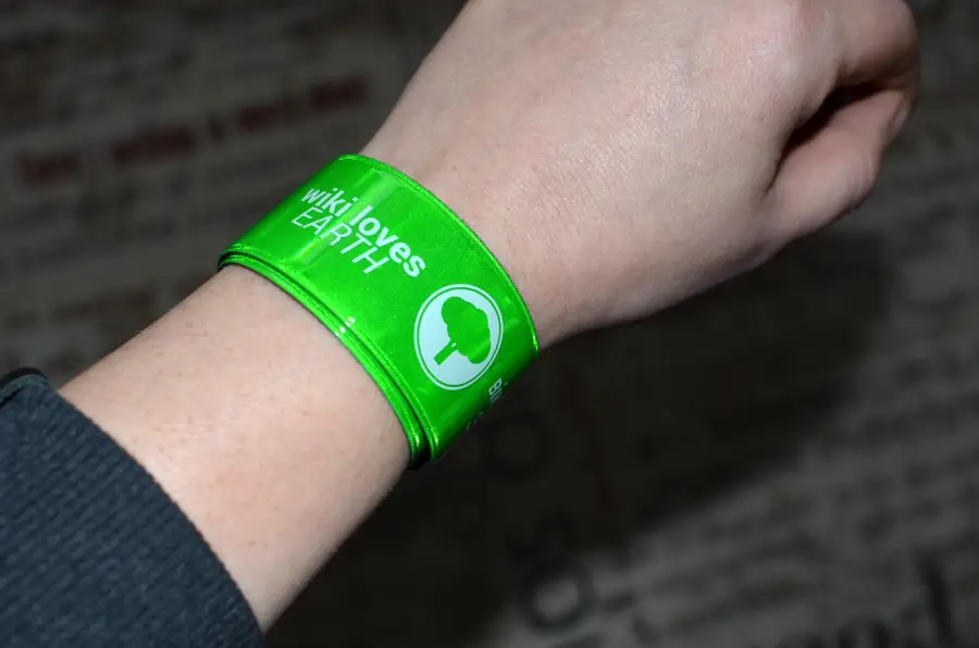 A person wearing a shiny green wristband with the text “wiki loves EARTH” and a white tree icon on their left wrist.