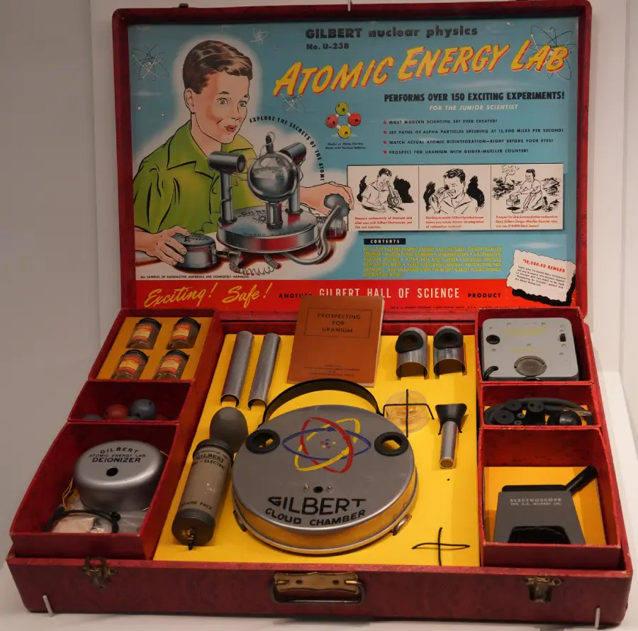 A vintage Atomic Energy Lab science kit with various components, including bottles, metal parts, and a cloud chamber, displayed in an open red case. The lid features colorful artwork and experiment instructions.