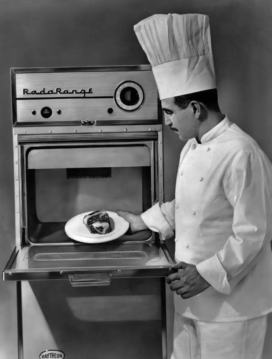 A chef in a tall hat places a plate with a cooked meal into an old-fashioned Raytheon Radarange microwave oven. The chef is wearing a white uniform and stands beside the open microwave door.
