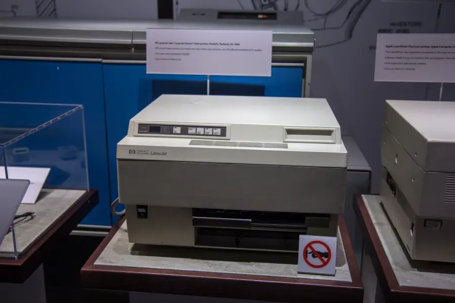 An HP LaserJet printer is displayed in a museum exhibit, with descriptive placards in the background and a no-touch sign in front of the printer.