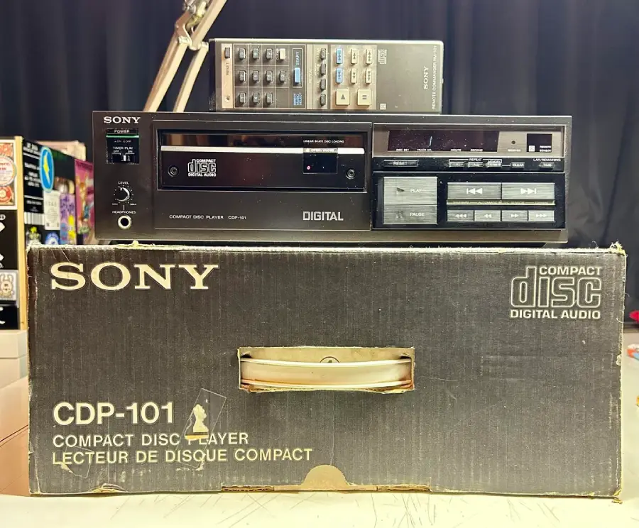 A vintage Sony CDP-101 compact disc player with remote control sits on top of its original black box. The box features SONY branding and product details in white text.