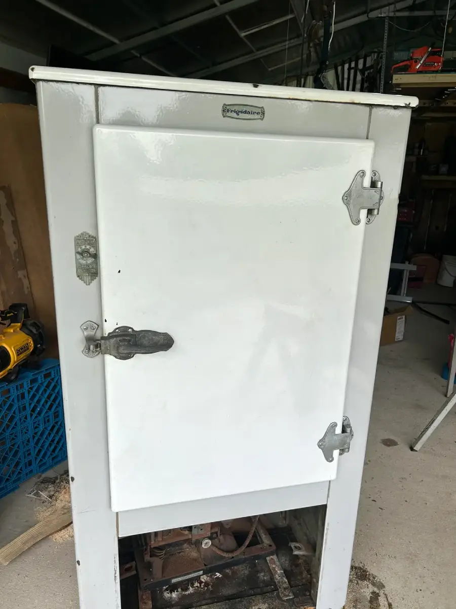 An antique white refrigerator with metal hinges and latches, standing in a garage with tools and boxes nearby. The brand name is visible on the top front of the fridge.