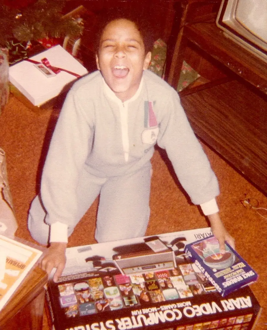 A child in pajamas excitedly kneels on the floor by an Atari Video Computer System and a Space Invaders game, smiling with mouth wide open. A wrapped gift and TV are visible in the background.