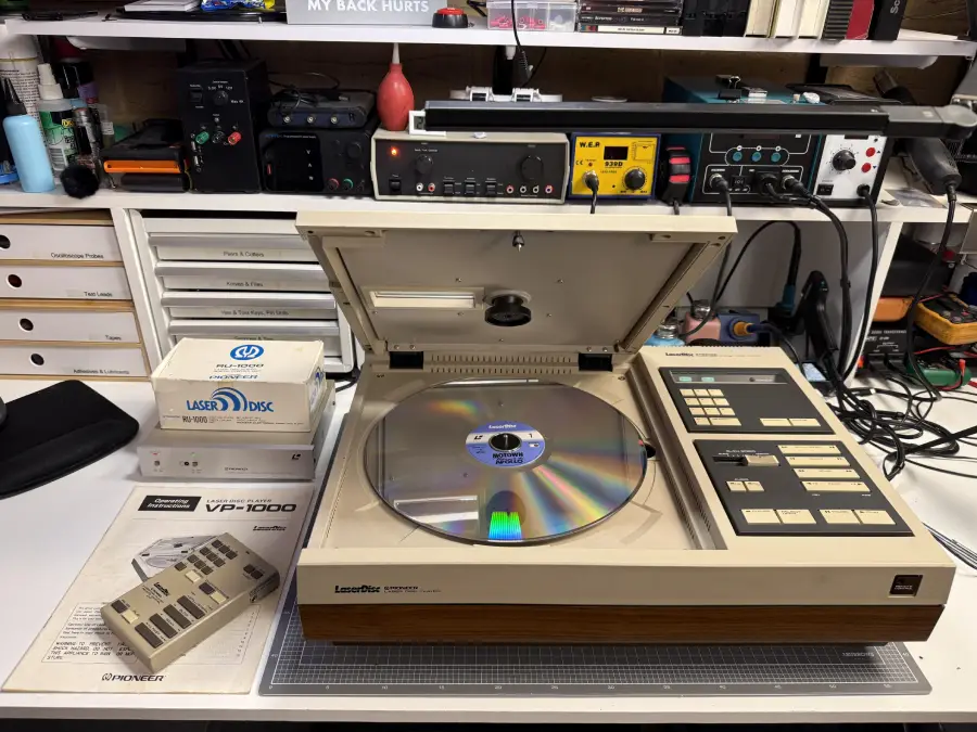 A vintage Pioneer LaserDisc player sits on a desk with its lid open, revealing a large disc inside. Nearby are a manual, remote, and LaserDisc packaging. Audio equipment and storage drawers are in the background.