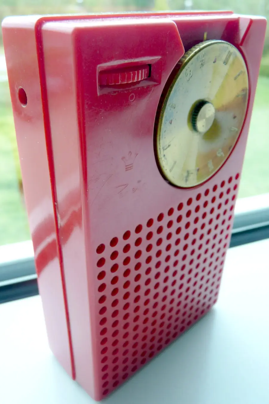 A close-up of a vintage red transistor radio with a round tuning dial and a small slider switch, resting on a windowsill.