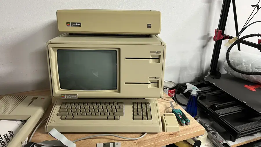 A vintage beige Apple Lisa computer with dual floppy drives, keyboard, and mouse sits on a desk beside electronics and tools in a workspace.