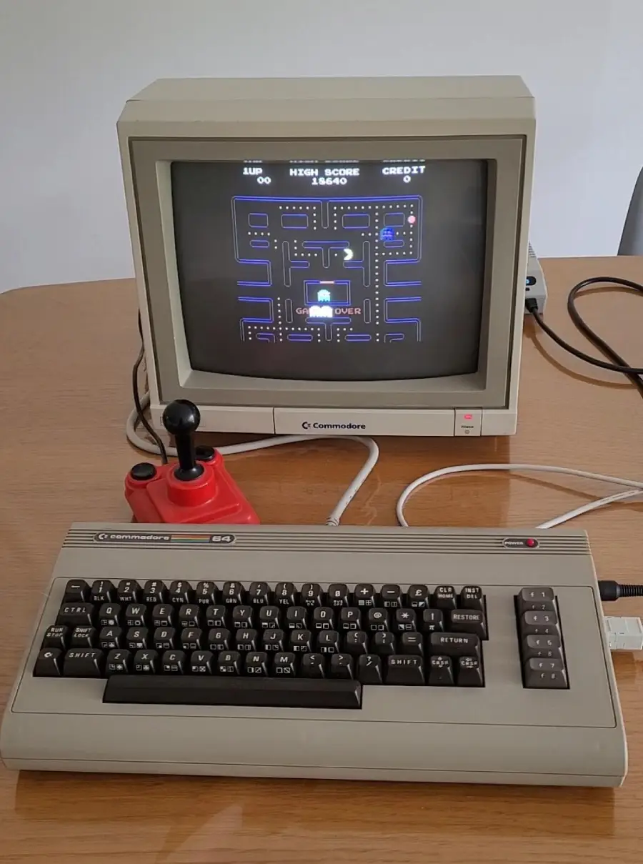 A vintage Commodore 64 computer setup with a monitor displaying a Pac-Man style game, a red joystick, and a beige keyboard on a wooden table.