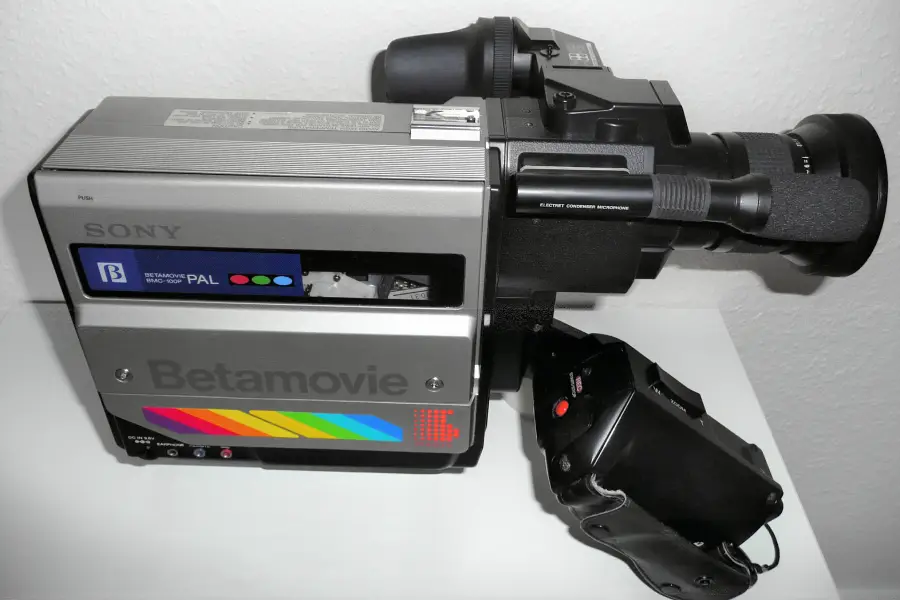 A vintage Sony Betamovie camcorder, featuring a silver body, Betamovie branding, and a rainbow stripe, sits on a white surface with a viewfinder and hand grip visible.