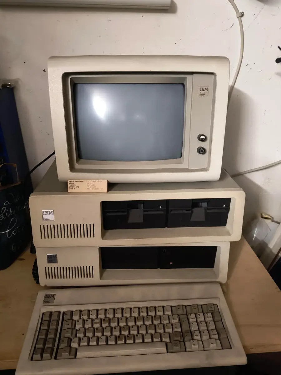 An IBM personal computer with a CRT monitor, two floppy disk drives, and a keyboard, all in beige, is sitting on a desk in a dimly lit room.
