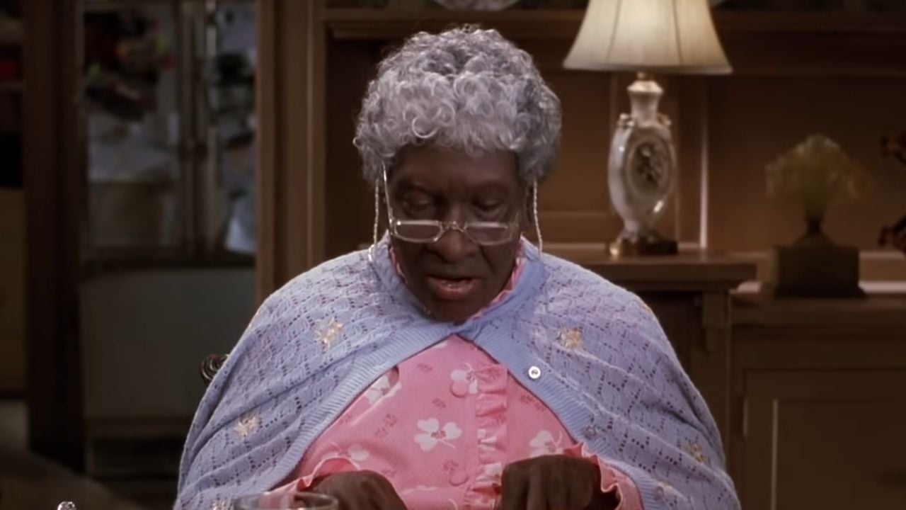 An elderly person with gray curly hair, glasses, and a blue shawl over a pink floral nightgown sits at a table reading, with a lamp and decorative items in the background.