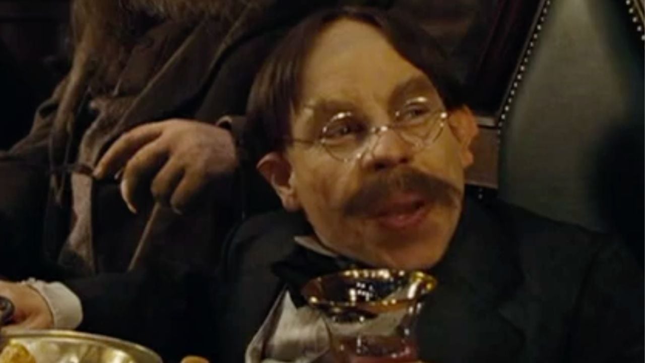 A man with glasses, a suit, and a mustache sits at a table holding a goblet, with food in front of him and another person partially visible beside him.