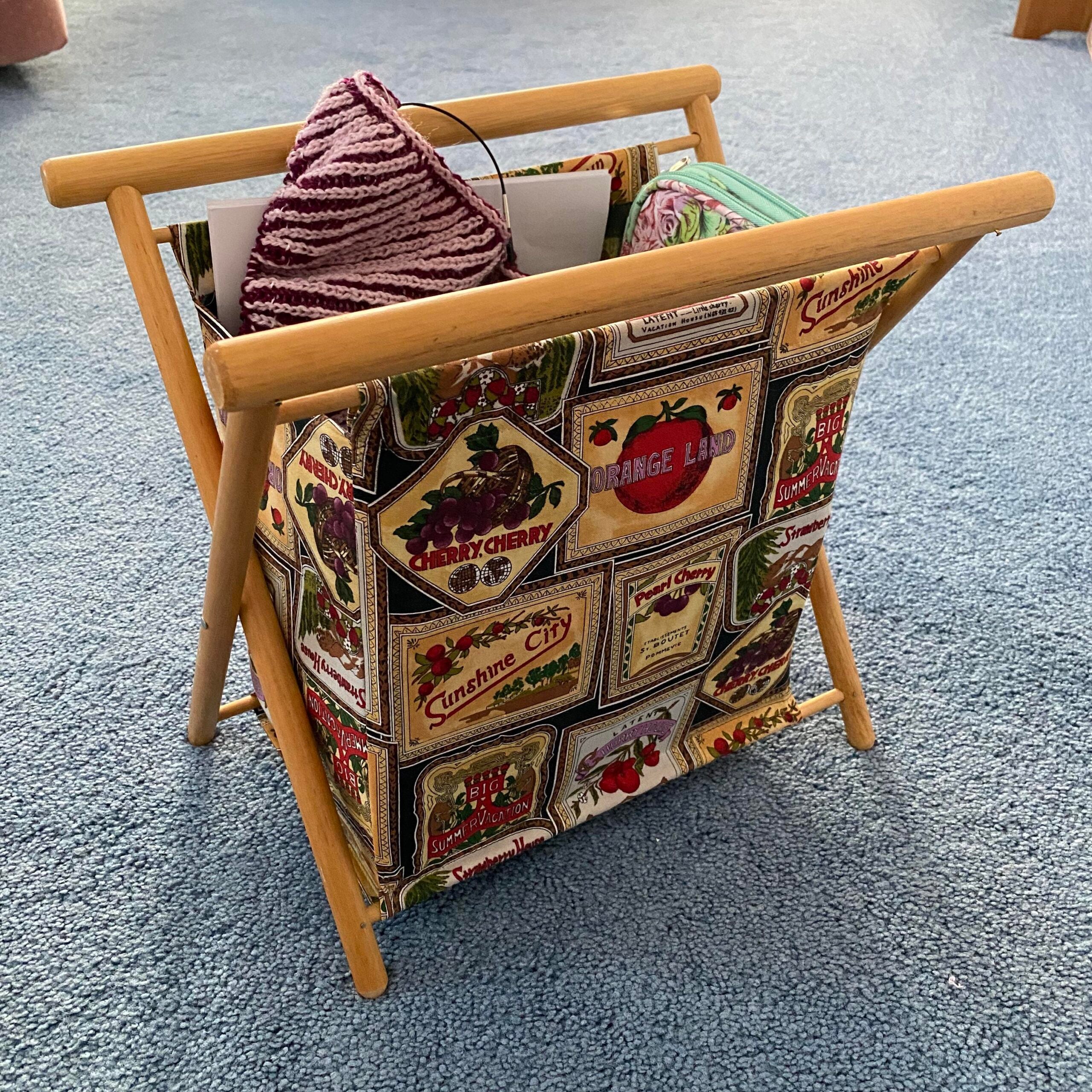 A wooden magazine rack with a vintage fruit label fabric holds yarn, a knitted item, and papers, sitting on blue carpet.