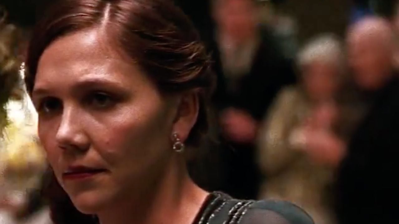 A woman with brown hair pulled back and wearing earrings looks off to the side with a serious expression at a formal event, with blurred people in the background.