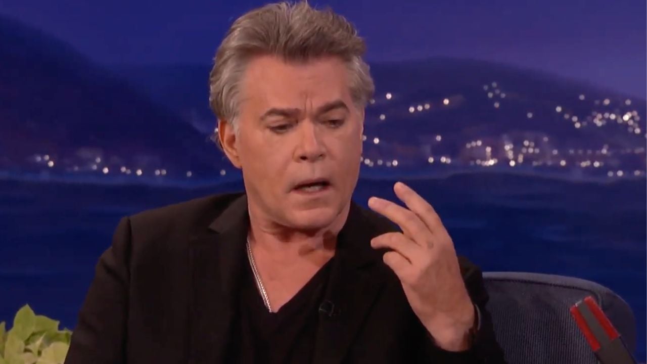 A man with gray hair in a black suit sits on a talk show set, holding up three fingers and looking at his hand, with a blurred cityscape in the background.