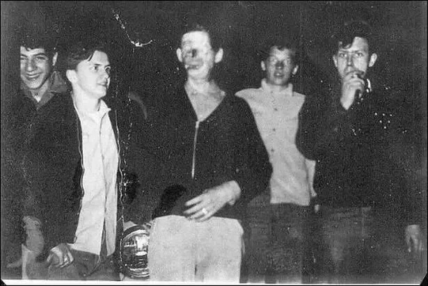 Black and white photo of five young men standing together at night, some smiling and one holding something to his mouth, with a dark background.