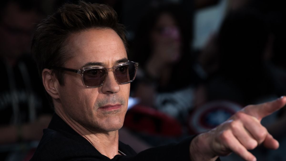A man wearing sunglasses and a dark jacket points forward with a serious expression. The background is blurred, showing people and bright lights.