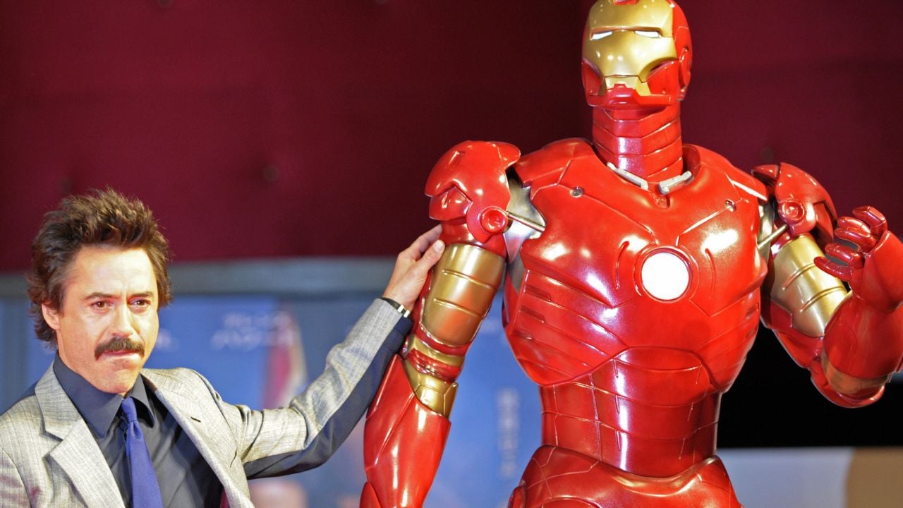 A man in a suit stands next to a life-sized Iron Man statue, resting his hand on its shoulder, with a red background behind them.