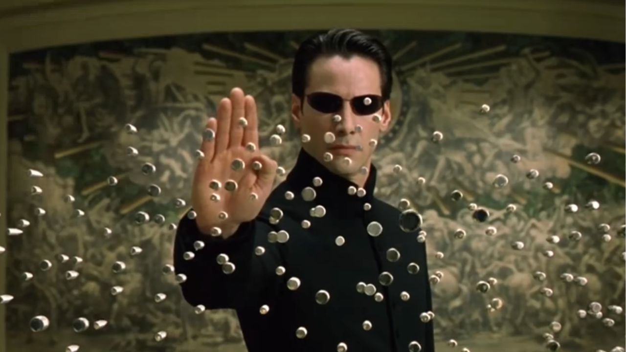 A man in black sunglasses and coat holds up his hand, stopping bullets mid-air in front of a detailed mural background.