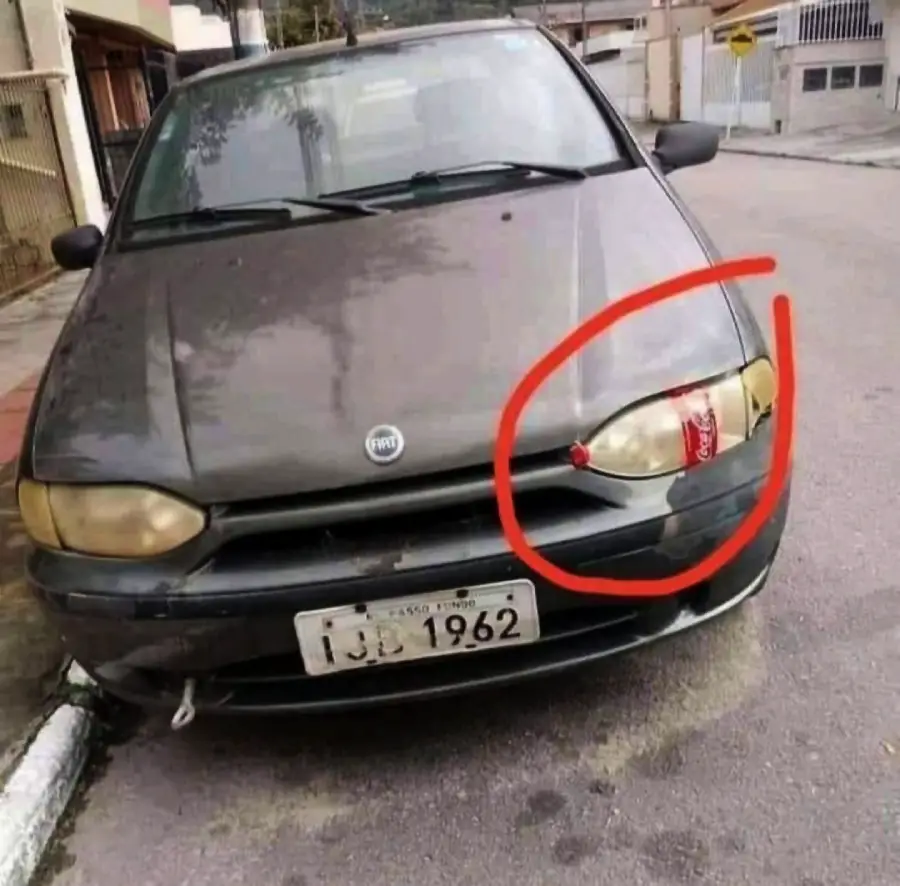 A dark car with one headlight replaced by a red Coca-Cola can, circled in red, parked on a quiet street.