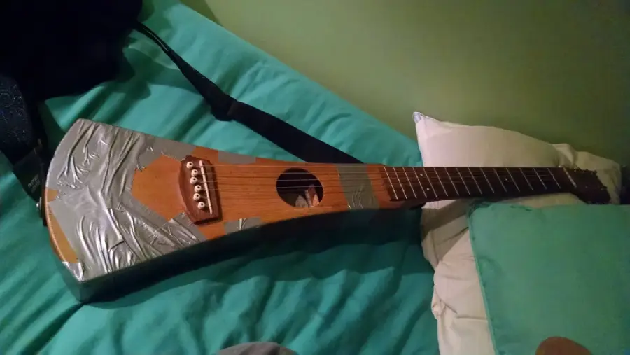 A wooden travel guitar with duct tape covering its lower body rests on a teal bedspread next to pillows.