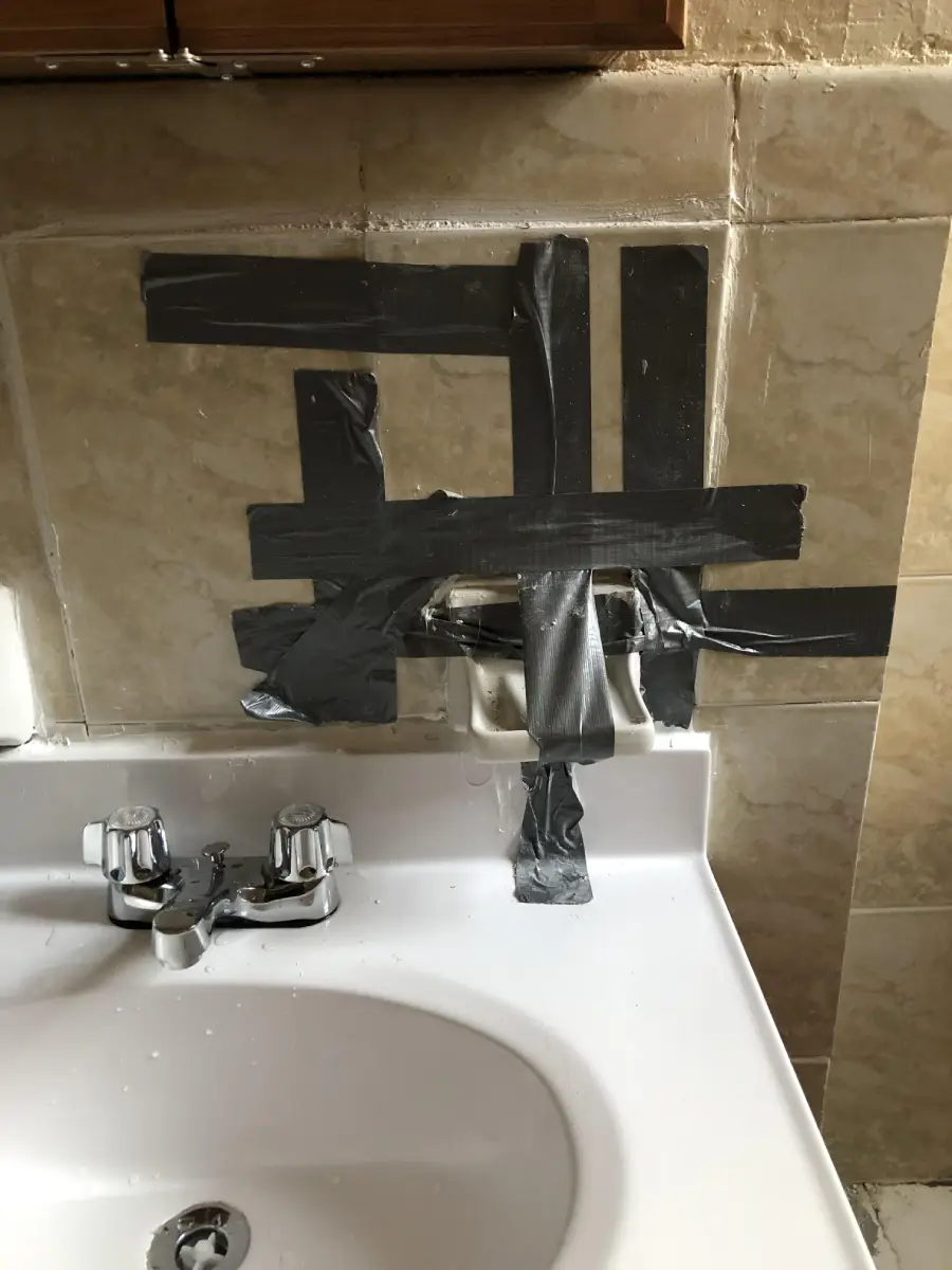 Bathroom sink with a faucet and surrounding wall patched up using strips of black duct tape in a makeshift repair, covering pipes and holes in beige tiles.