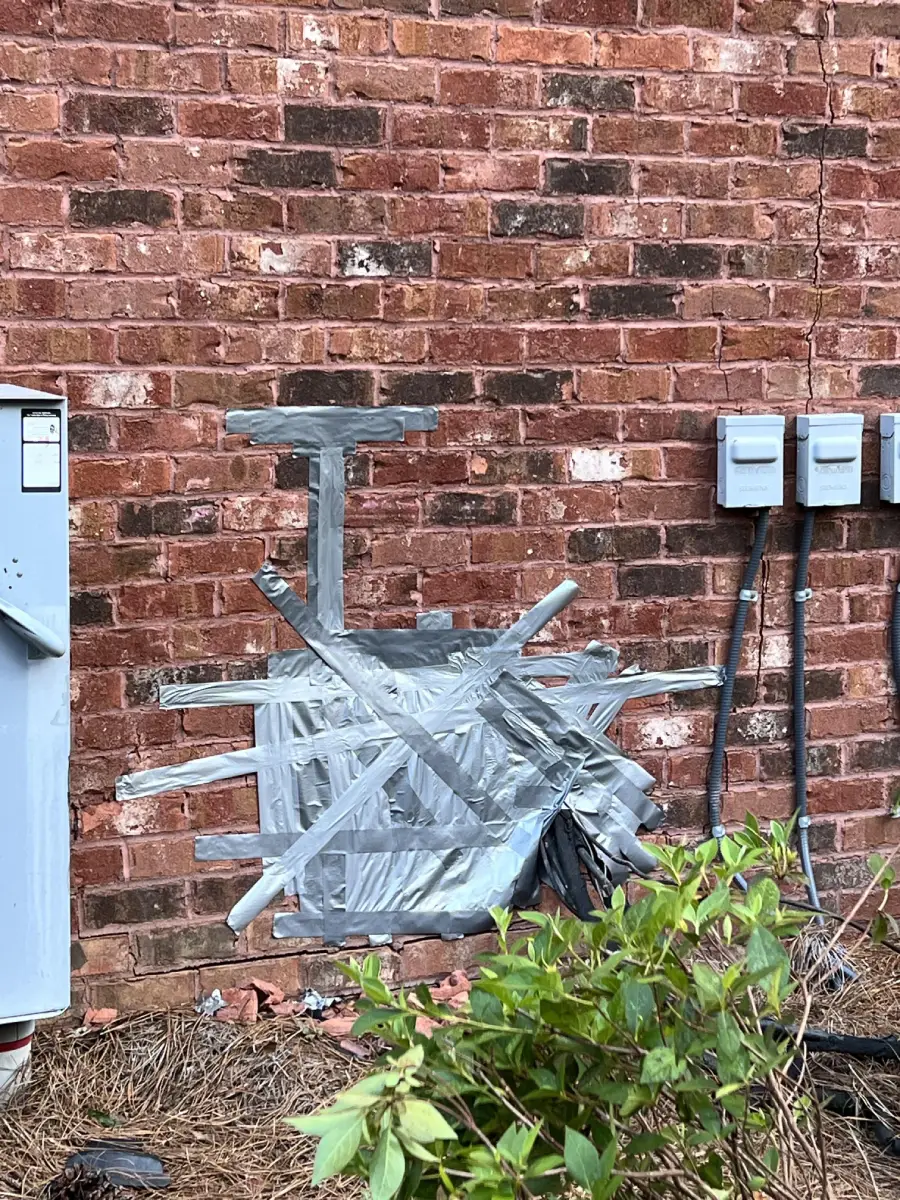 An outdoor air conditioning unit is heavily wrapped in silver duct tape and attached to a brick wall. Electrical meters and a green bush are nearby.