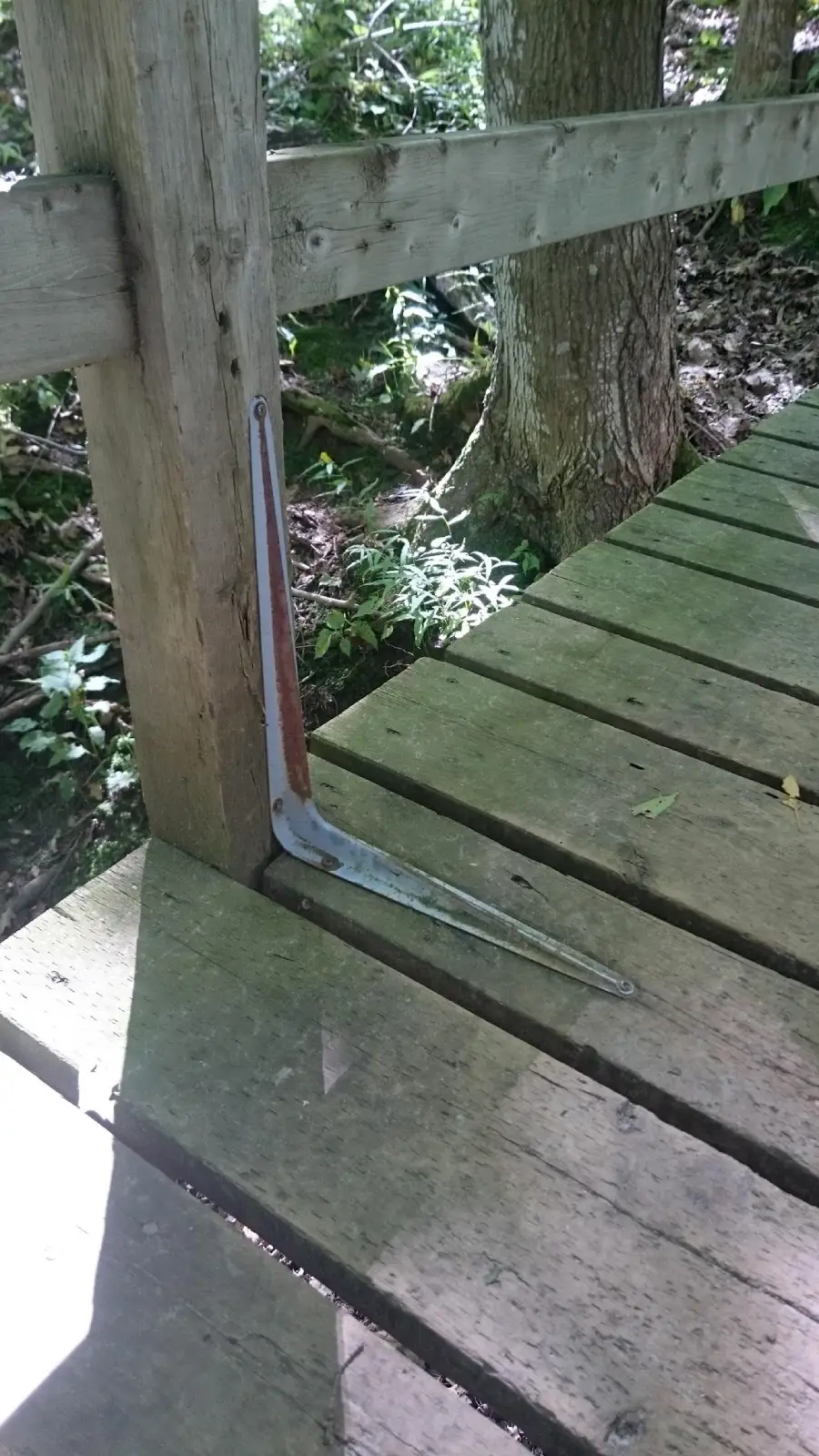 A metal bracket is attached to a wooden post and deck. Sunlight casts shadows across the wooden planks, with green foliage and part of a tree trunk visible in the background.