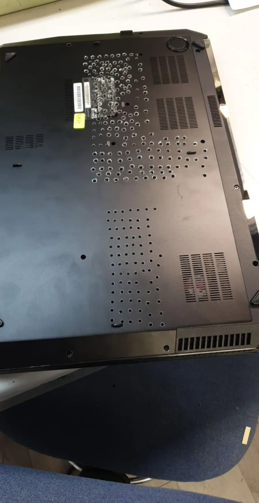 The image shows the bottom of a black laptop with many holes drilled into its case, likely for ventilation. The laptop rests on a chair with part of a workspace visible in the background.