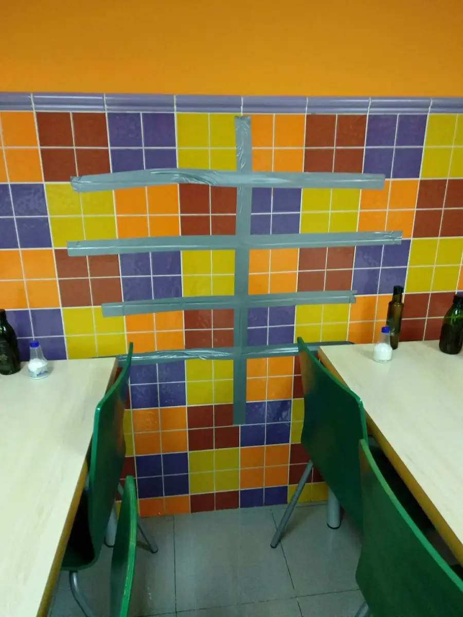 Four silver duct tape strips form a hashtag on a colorful tiled wall between two tables with green chairs. Several bottles and salt or pepper shakers are on the tables.