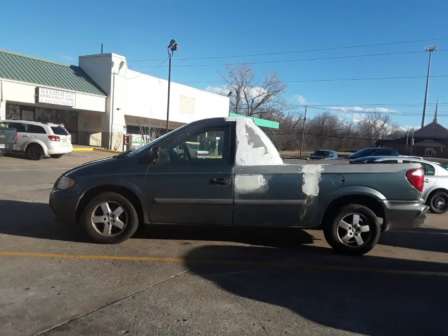 A minivan modified to resemble a pickup truck, with visible signs of white patchwork and bodywork, is parked in a lot on a sunny day. Other cars and shops are in the background.