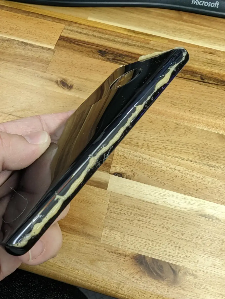 A hand holds a smartphone with its back cover partially detached and yellow adhesive oozing out along the seam. The phone is on a wooden surface.