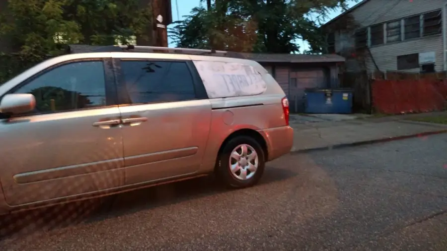 A silver minivan is parked on a street near a house with boarded windows and a blue dumpster. The photo appears to be taken at dusk.